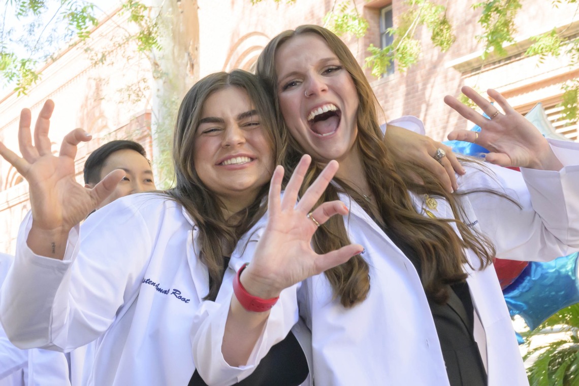 Two University of Arizona R. Ken Coit College of Pharmacy students smile and make the Wildcat sign with their fingers as they walk outside after receiving their white coats.