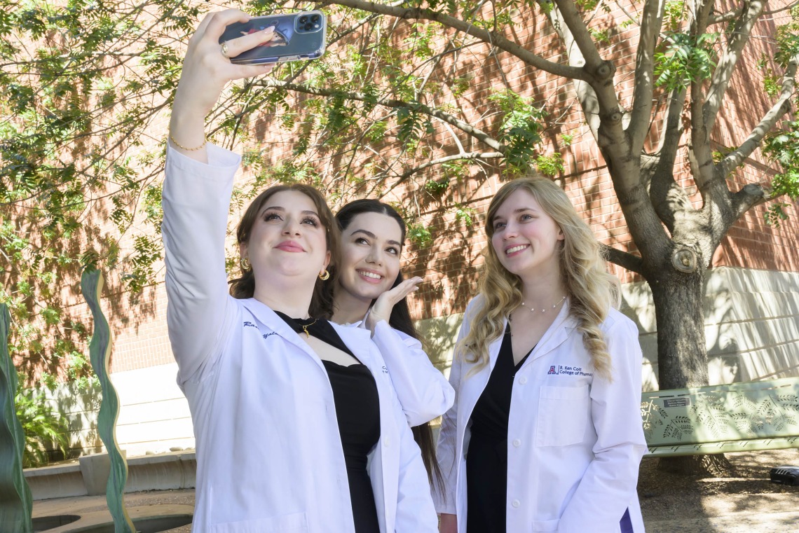 Three University of Arizona R. Ken Coit Pharmacy students in white coats pose for a selfie in an outdoor space. 