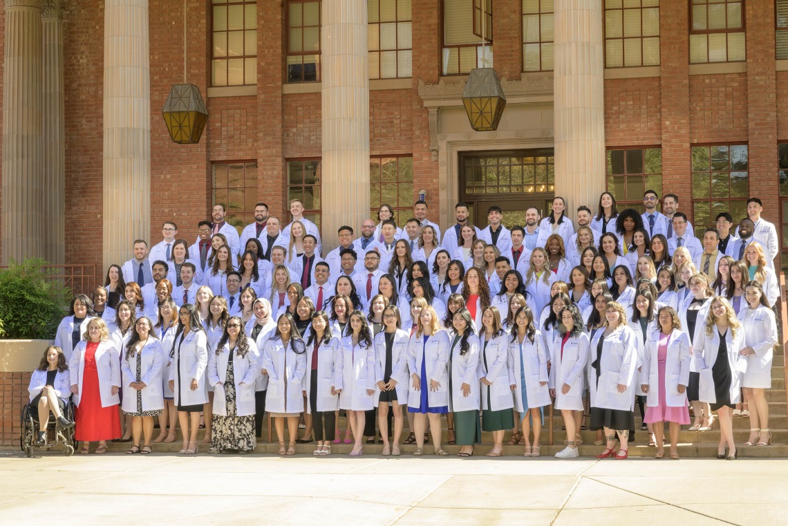 128 University of Arizona R. Ken Coit College of Pharmacy third-year students gather for a group photo on the steps outside a building wearing their white coats. 