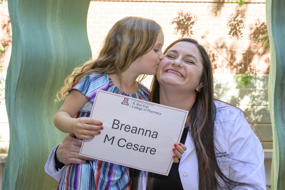 A young girl kisses her mom’s cheek while holding a sign that says “Breanna M. Cesare”. 