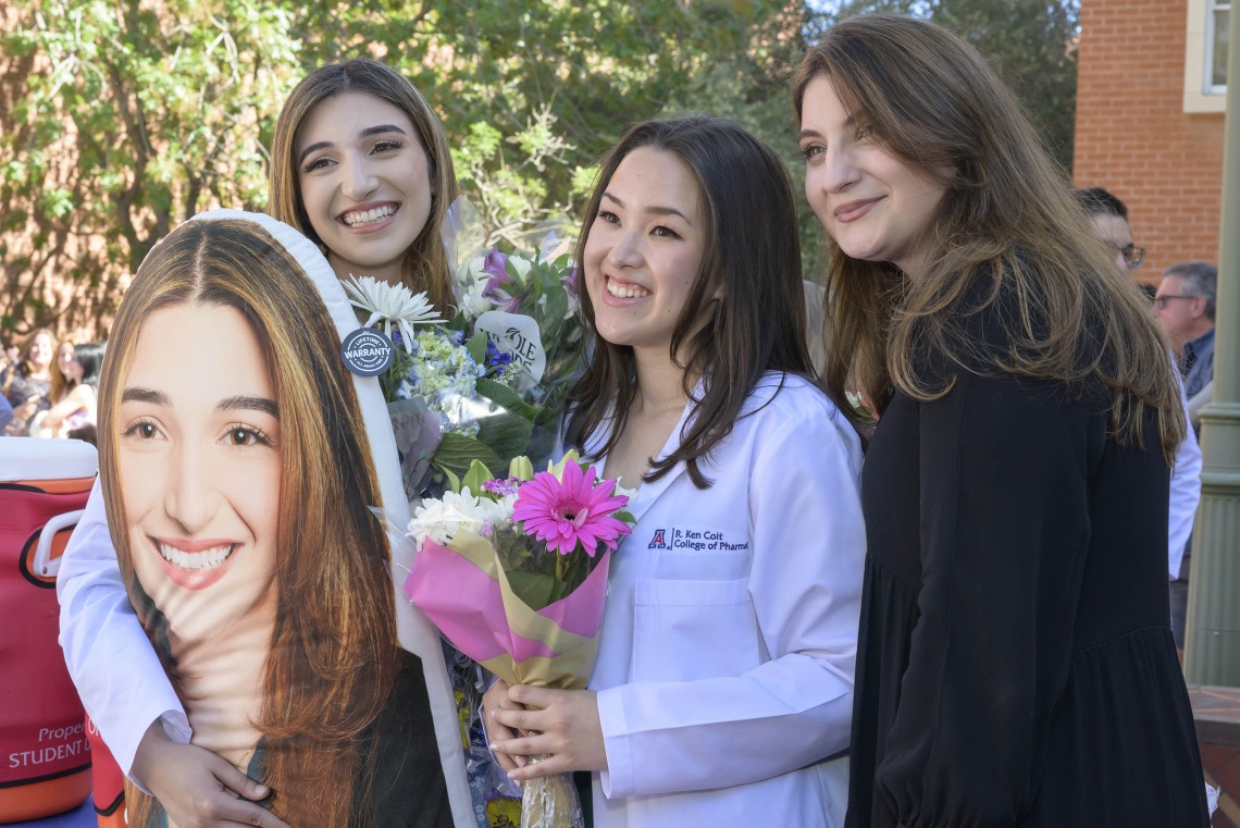 Two University of Arizona R. Ken Coit College of Pharmacy students and one graduate stand together with flowers and a large photo. 