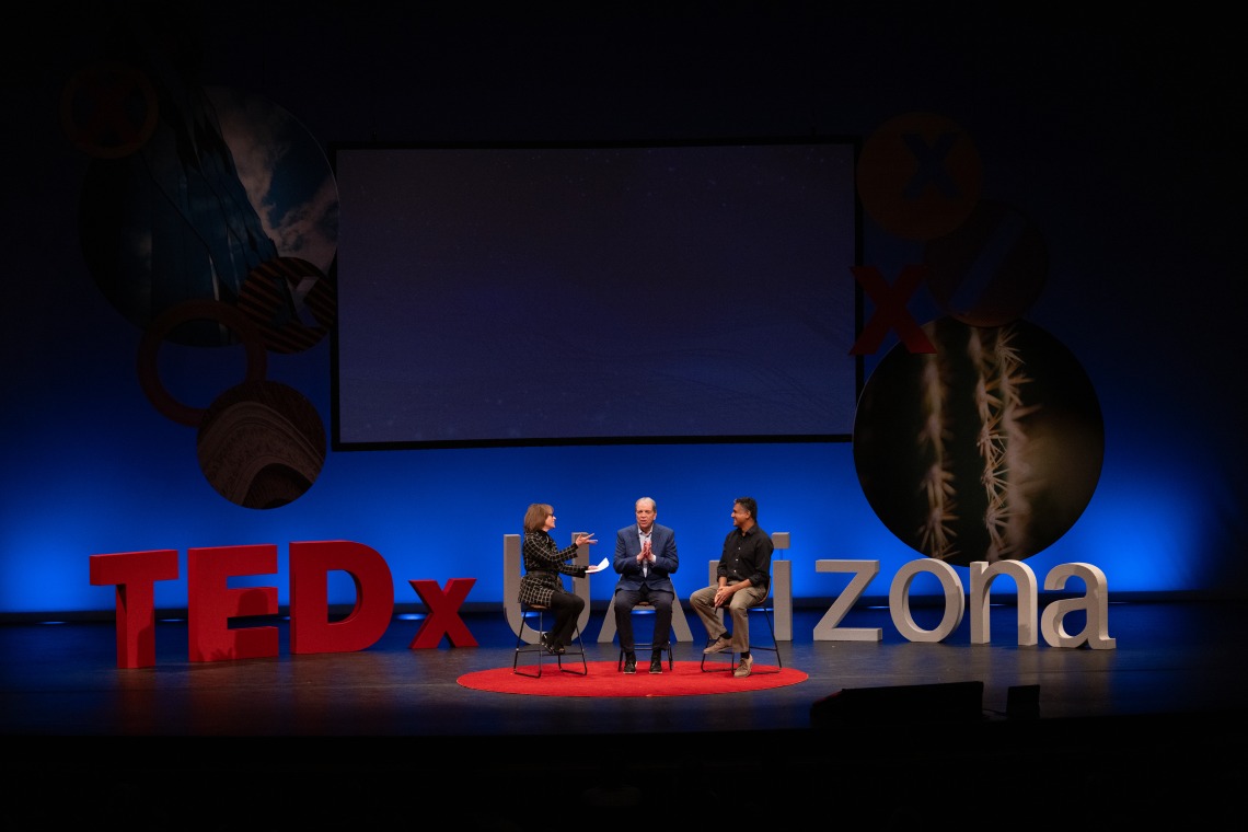 Three people sit on a stage in front of a large sign that says “UArizona.”