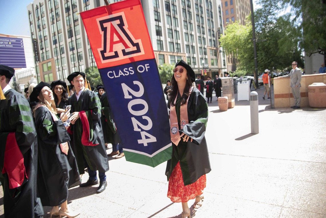 A University of Arizona College of Medicine – Phoenix student dressed in a graduation cap and gown carries a large banner that says “Class of 2024” as she walks outside. 