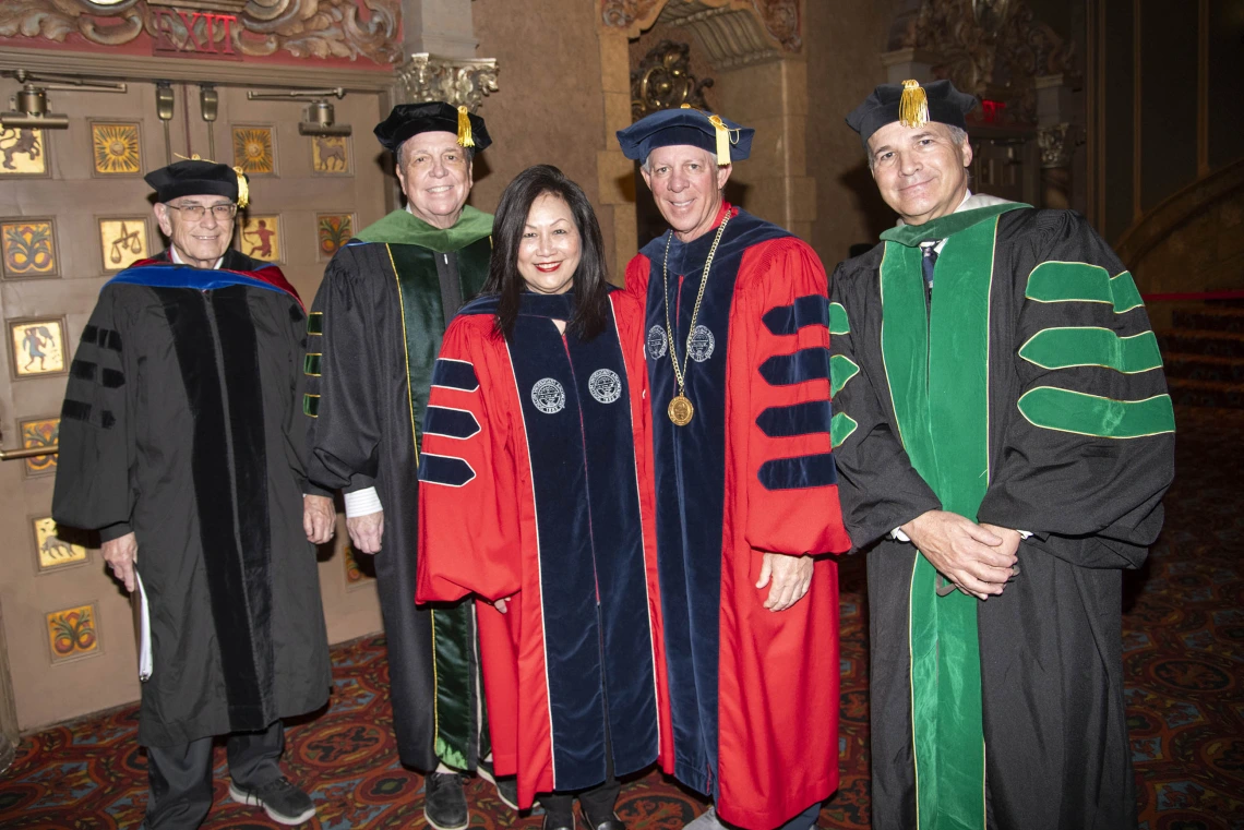 Five people dressed in graduation regalia stand together smiling. 