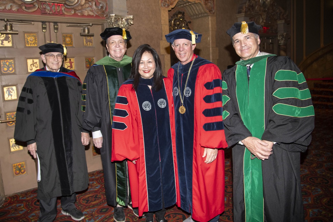 Five people dressed in graduation regalia stand together smiling. 