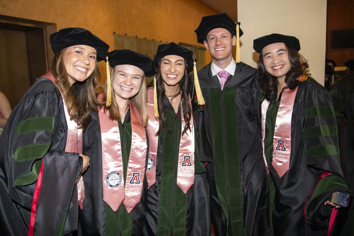 Five University of Arizona College of Medicine – Phoenix students dressed in graduation regalia stand together smiling before their commencement ceremony.