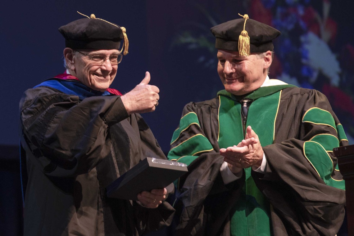 Two professors dressed in graduation regalia stand next to each other smiling as one hods a plaque and gives a thumbs-up. 
