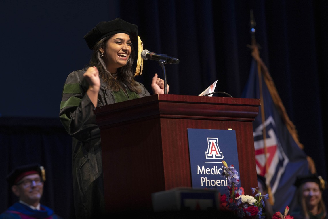 A University of Arizona College of Medicine – Phoenix student dressed in graduation regalia stands at a podium, smiling and gesturing with her hands, during the commencement ceremony.