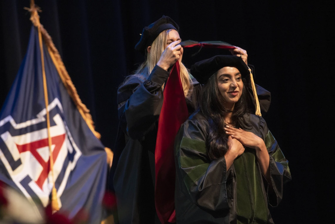 A University of Arizona College of Medicine – Phoenix student in graduation regalia holds her hands over her heart as a professor places a graduation hood on her.