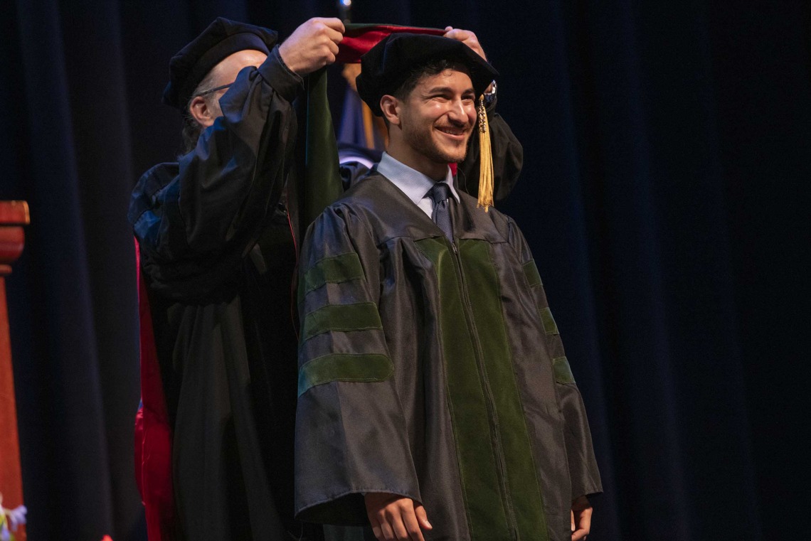 A University of Arizona College of Medicine – Phoenix student in graduation regalia smiles as a professor places a graduation hood on him. 