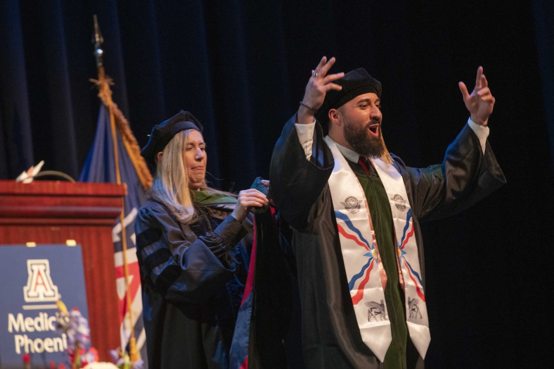 A University of Arizona College of Medicine – Phoenix student in graduation regalia holds his hands in the air in celebration as a professor standing behind him prepares to place a graduation hood on him.