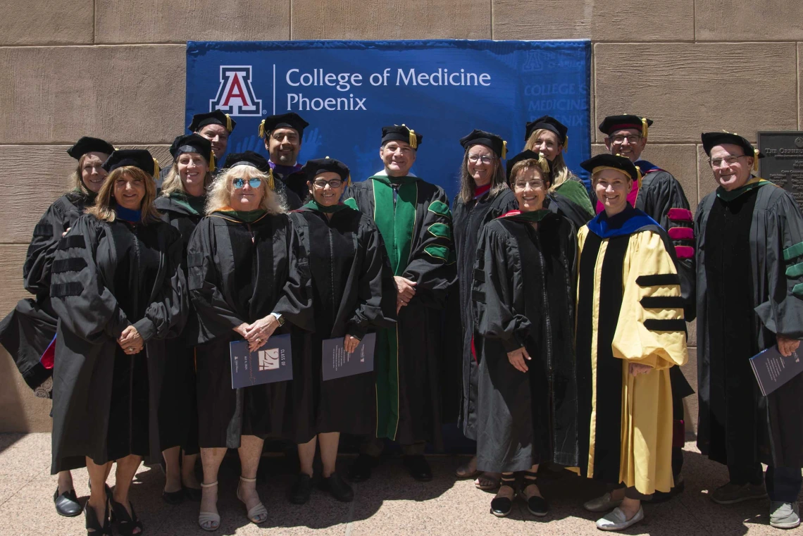 Over a dozen University of Arizona College of Medicine – Phoenix faculty members wearing graduation regalia stand in front of a sign that reads, “College of Medicine – Phoenix.”