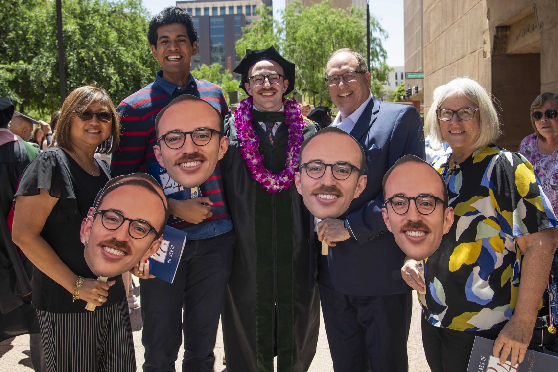 Several family members holding large faces on sticks surround a newly graduated medical student wearing a cap and gown after the University of Arizona College of Medicine – Phoenix commencement. 