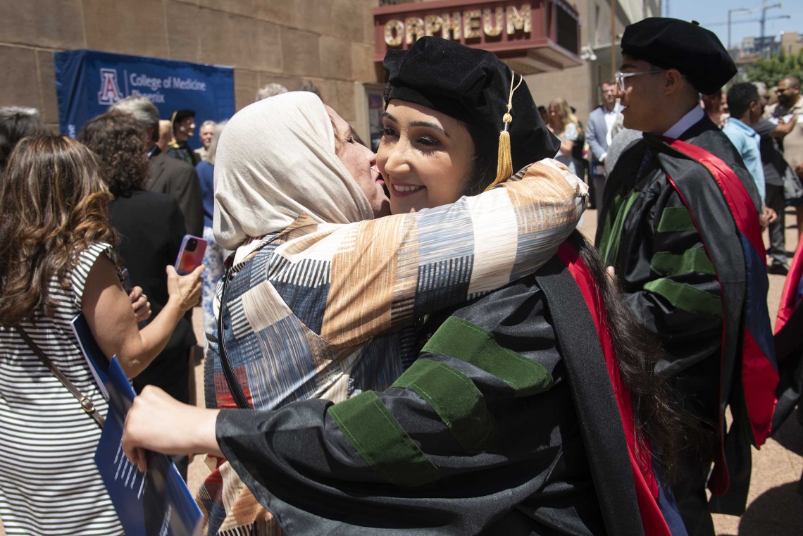 A woman in a hijab hugs her daughter, who is wearing graduation regalia outside amid a crowd of people.