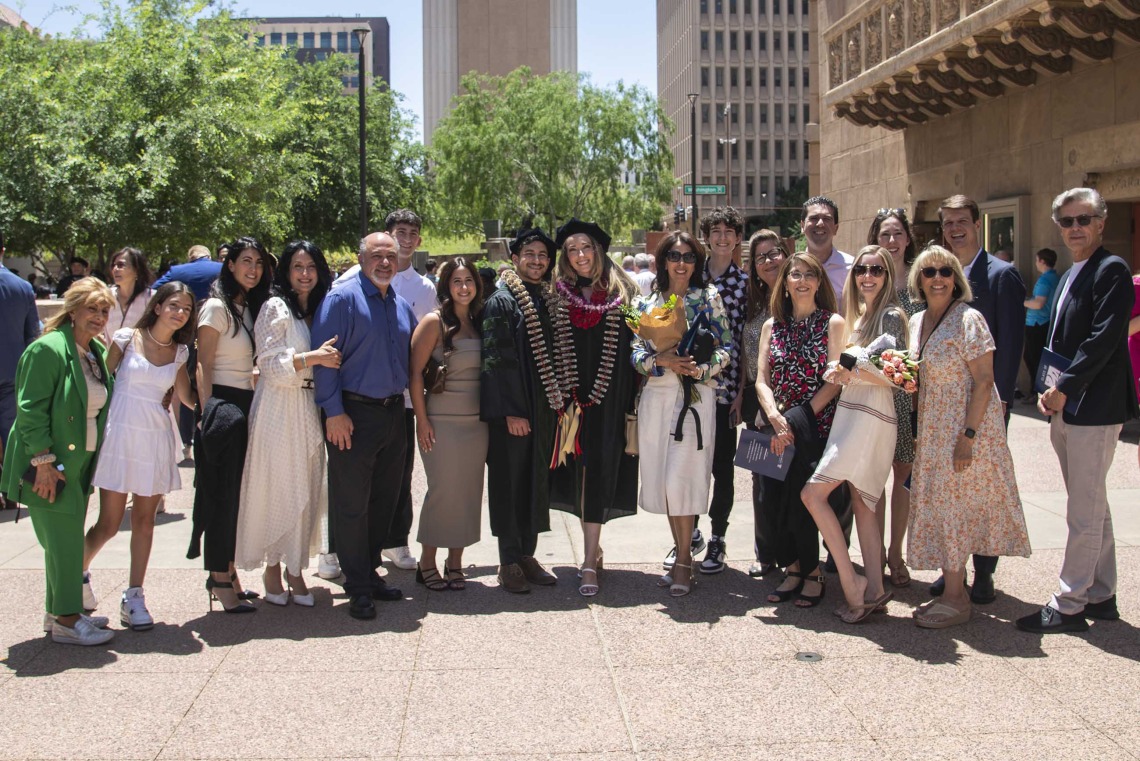 A large group of family members stand on each side of two University of Arizona College of Medicine – Phoenix graduates wearing their caps and gowns. 