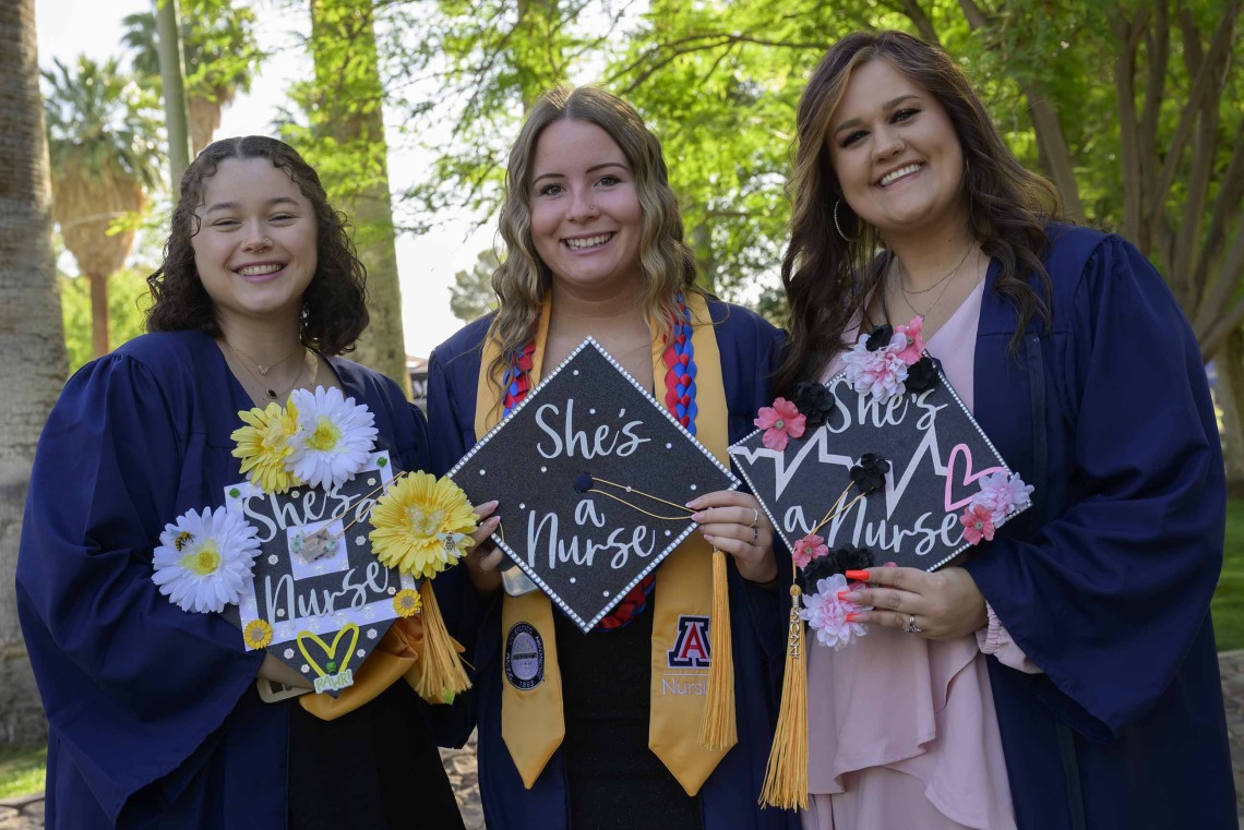 Three University of Arizona College of Nursing Bachelor of Science in Nursing students in graduation gowns show the decorated tops of their graduation caps. 