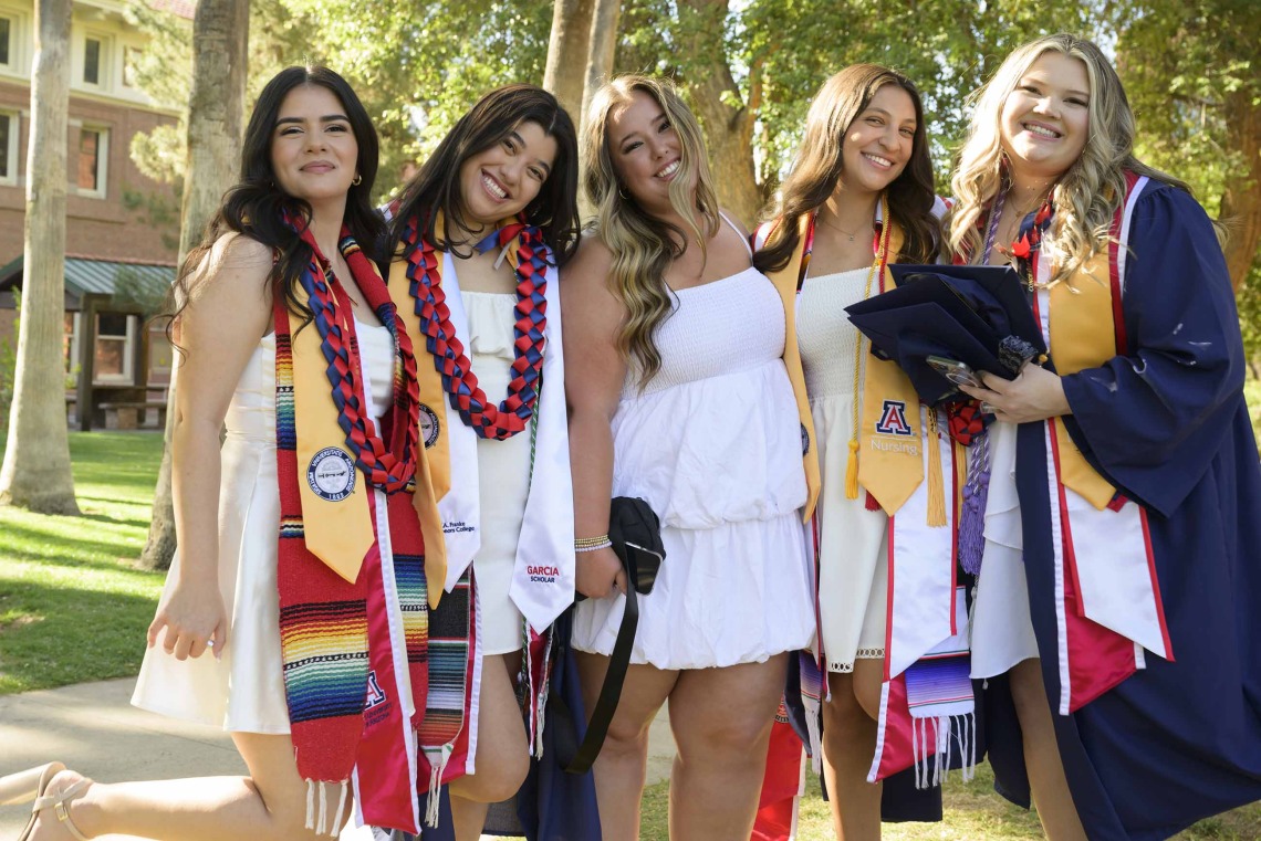 Five University of Arizona College of Nursing Bachelor of Science in Nursing students smile as they stand together outside. Some are wearing graduation gowns and have University of Arizona sashes on. 