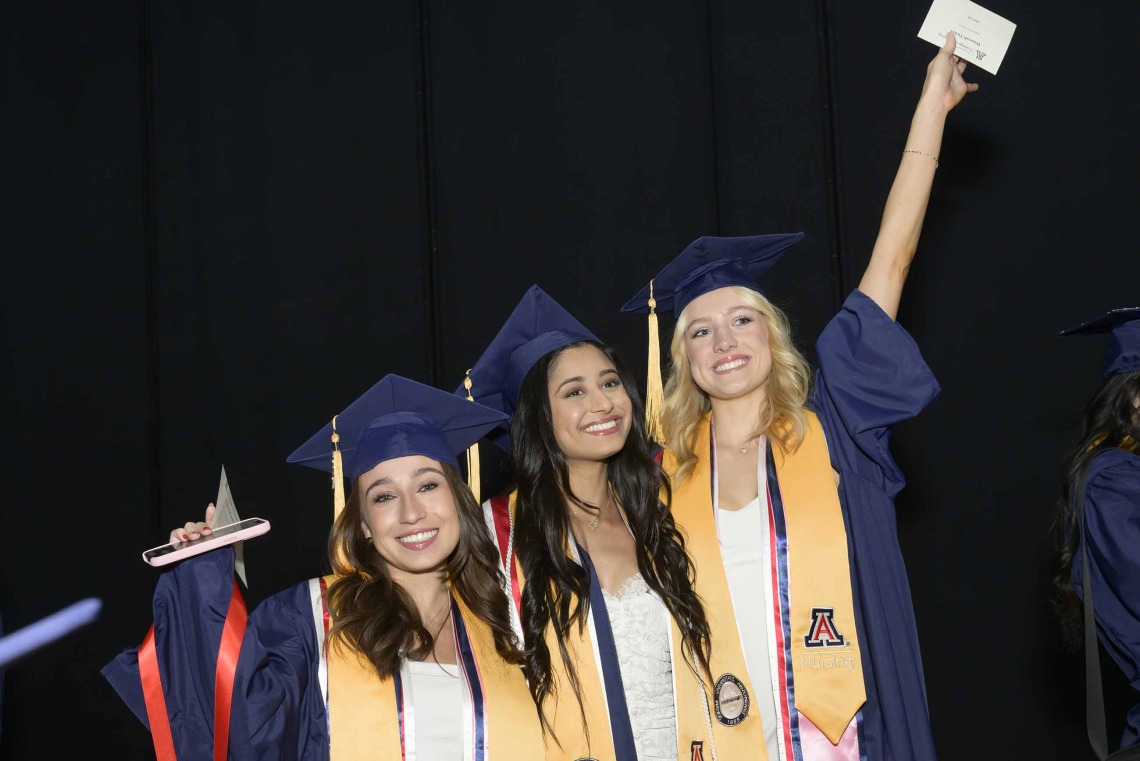 Three University of Arizona College of Nursing students in graduation regalia smile and raise hands in the air in celebration of their graduation.  
