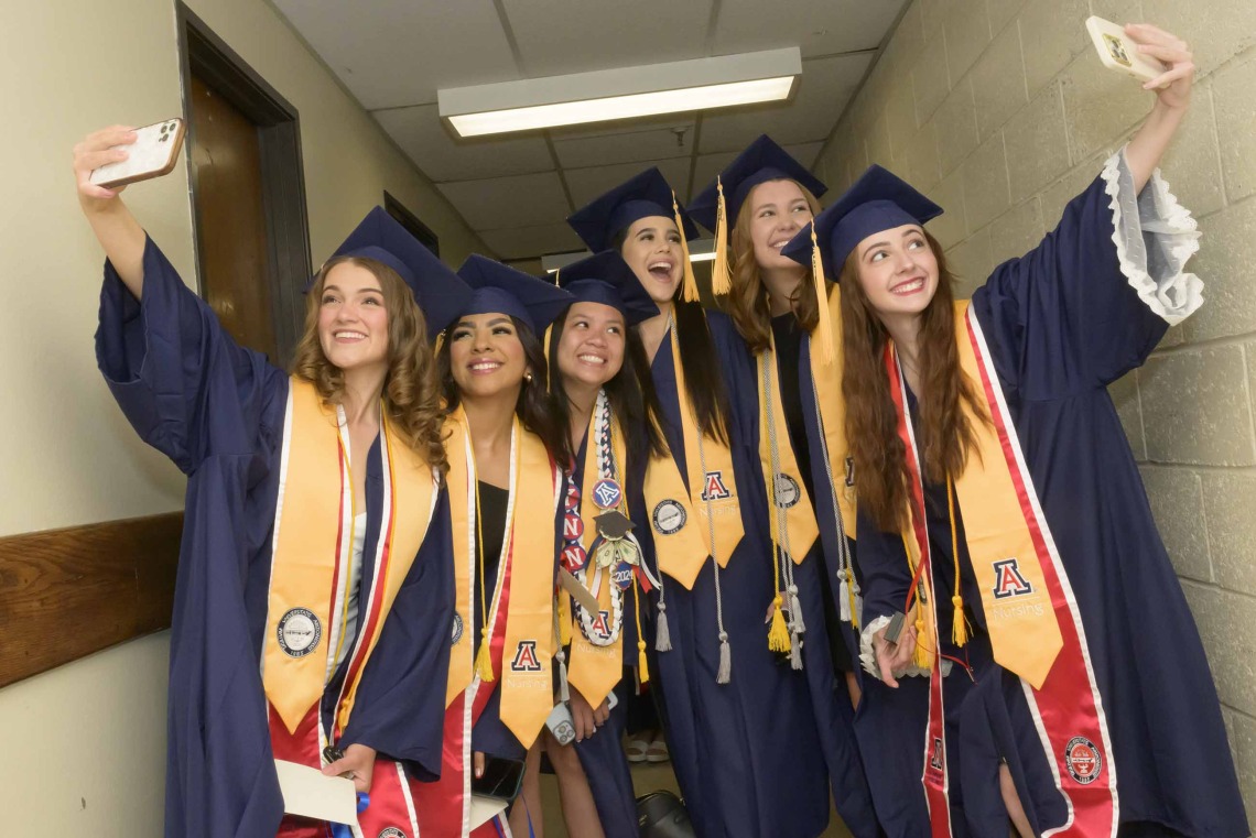 Six University of Arizona College of Nursing students dressed in graduation caps and gowns stand in a hallway while two of them are taking selfies. 