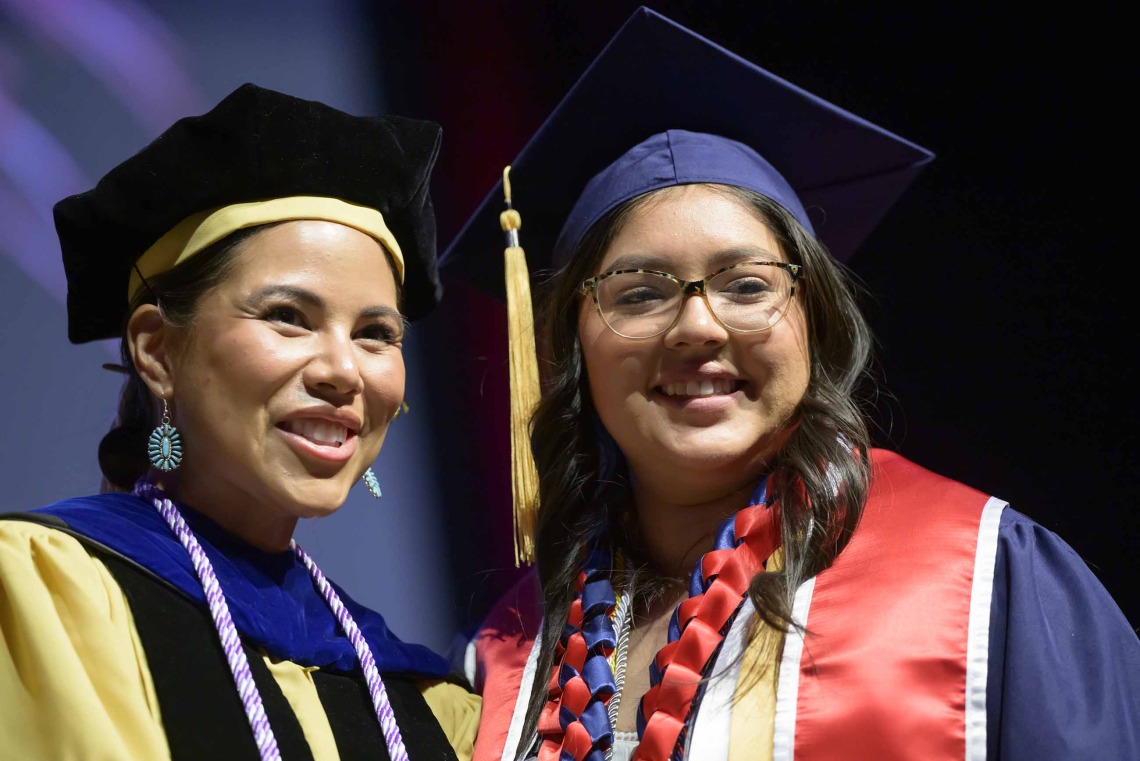 A University of Arizona College of Nursing professor and student, both wearing graduation caps and gowns, stand side by side smiling.