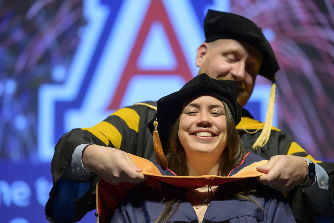 A University of Arizona College of Nursing professor places a hood over the shoulders of a nursing student. Both are dressed in graduation caps and gowns.