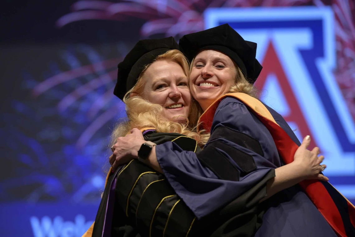 A University of Arizona College of Nursing professor hugs a nursing student. Both are dressed in graduation caps and gowns.