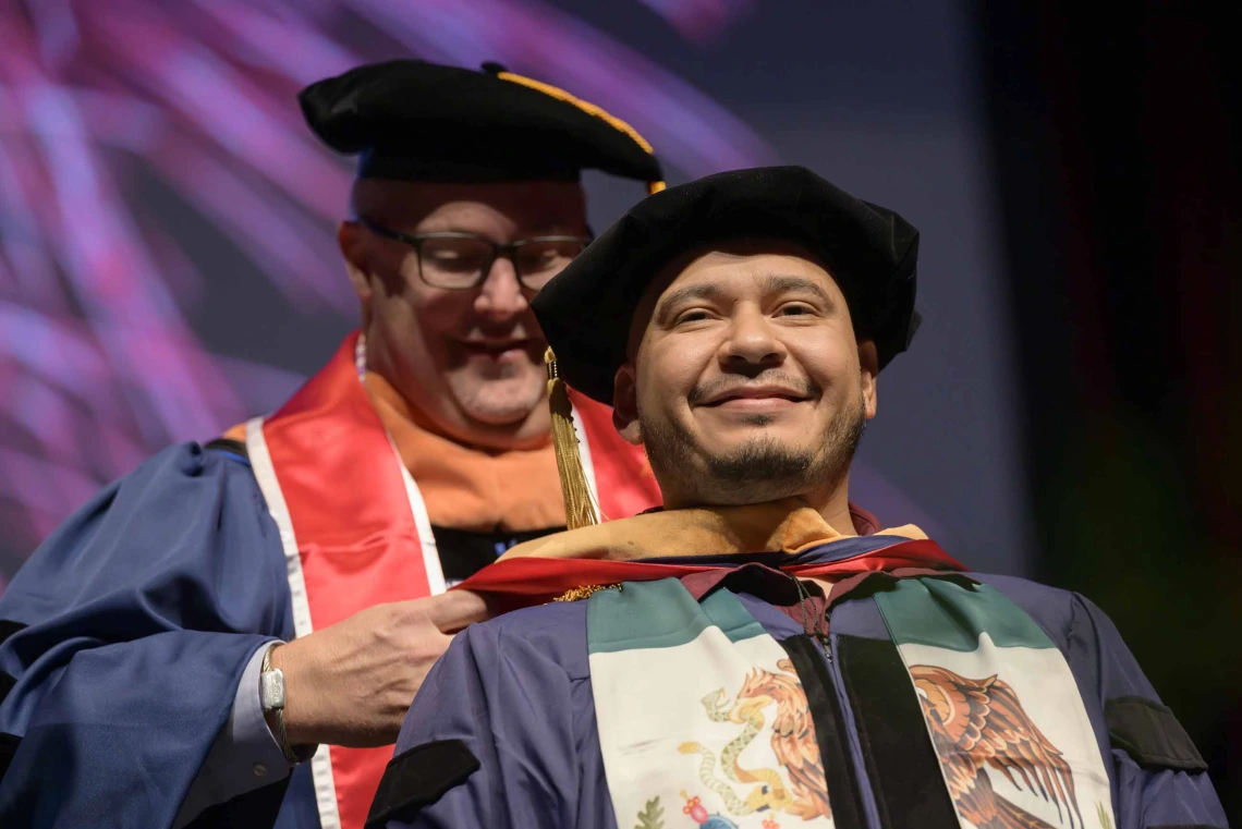 A University of Arizona College of Nursing professor places a hood over the shoulders of a nursing student. Both are dressed in graduation caps and gowns. 