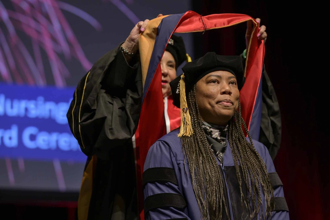 A University of Arizona College of Nursing professor places a hood over the shoulders of a nursing student. Both are dressed in graduation caps and gowns.