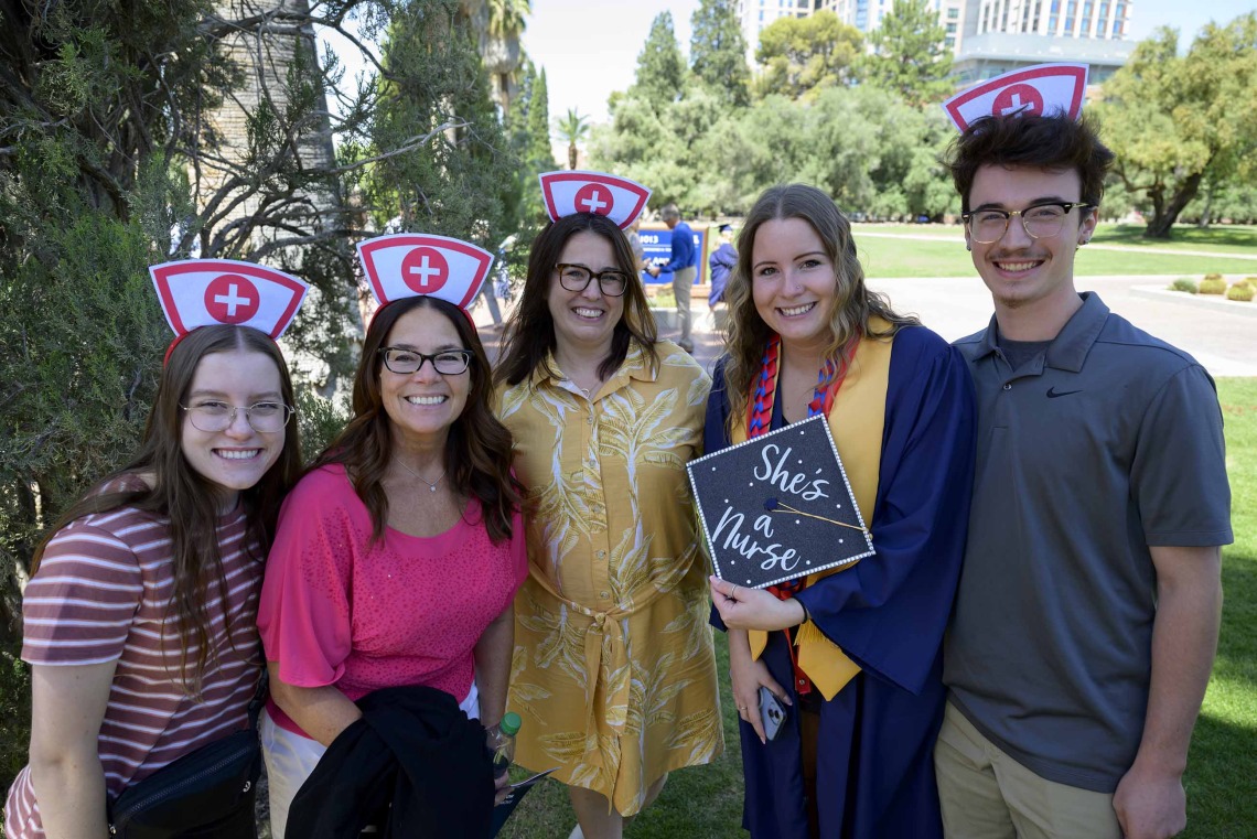 A family stands on either side of a recent graduate of the University of Arizona College of Nursing who is dressed in a graduation cap and gown. The family members have white paper hats with a red cross on them. 