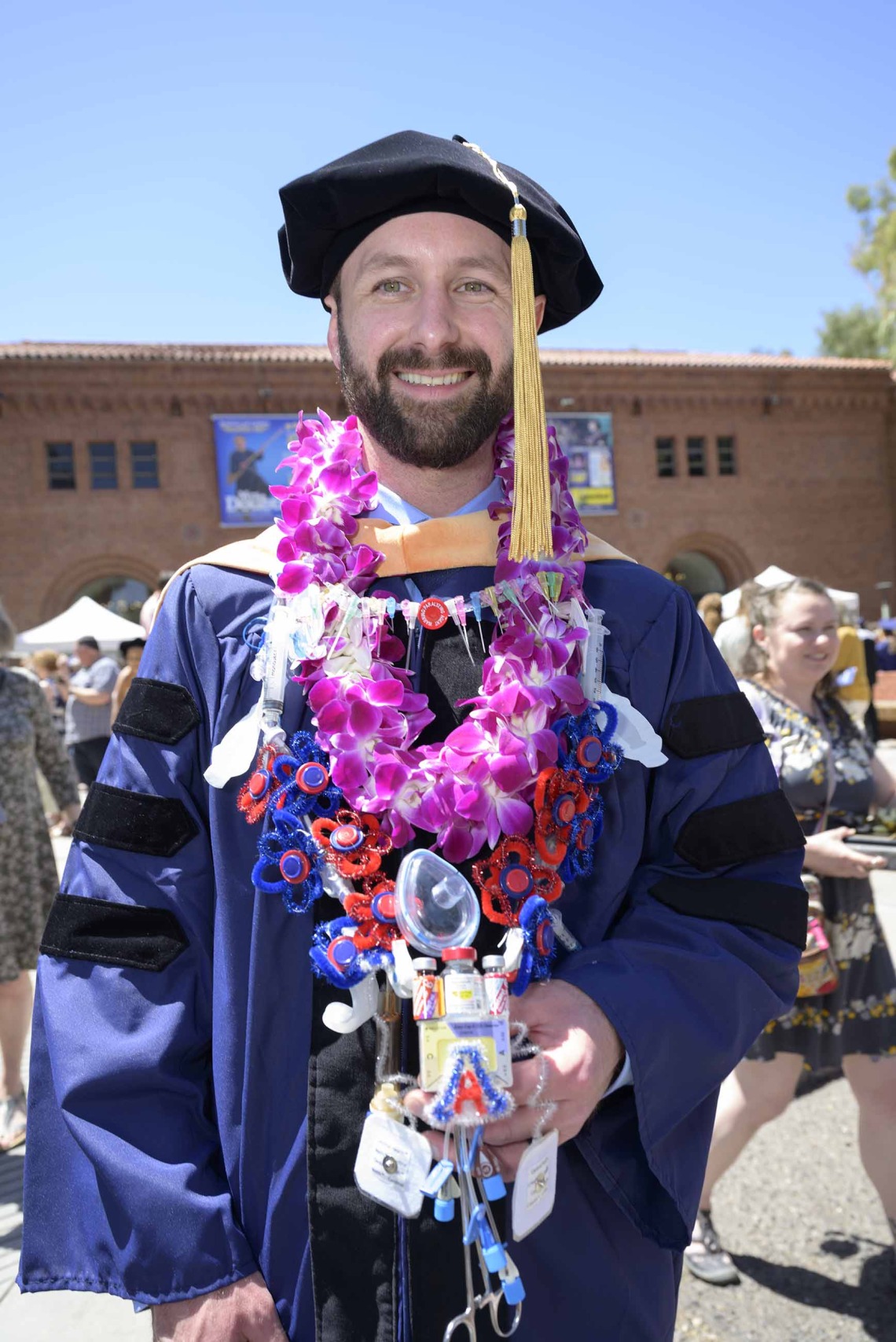A University of Arizona College of Nursing doctor of nursing practice graduate stands outside smiling in a graduation cap and gown. He has flowers around his neck and a necklace made out of a variety of medical supplies used in his profession. 