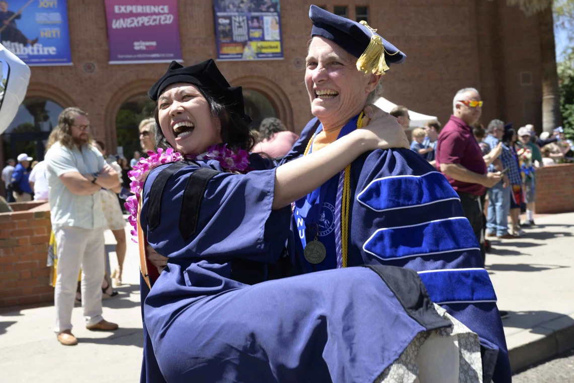 A University of Arizona College of Nursing professor carries a Nurse-Midwifery graduate. Both are dressed in graduation caps and gowns and laughing. 