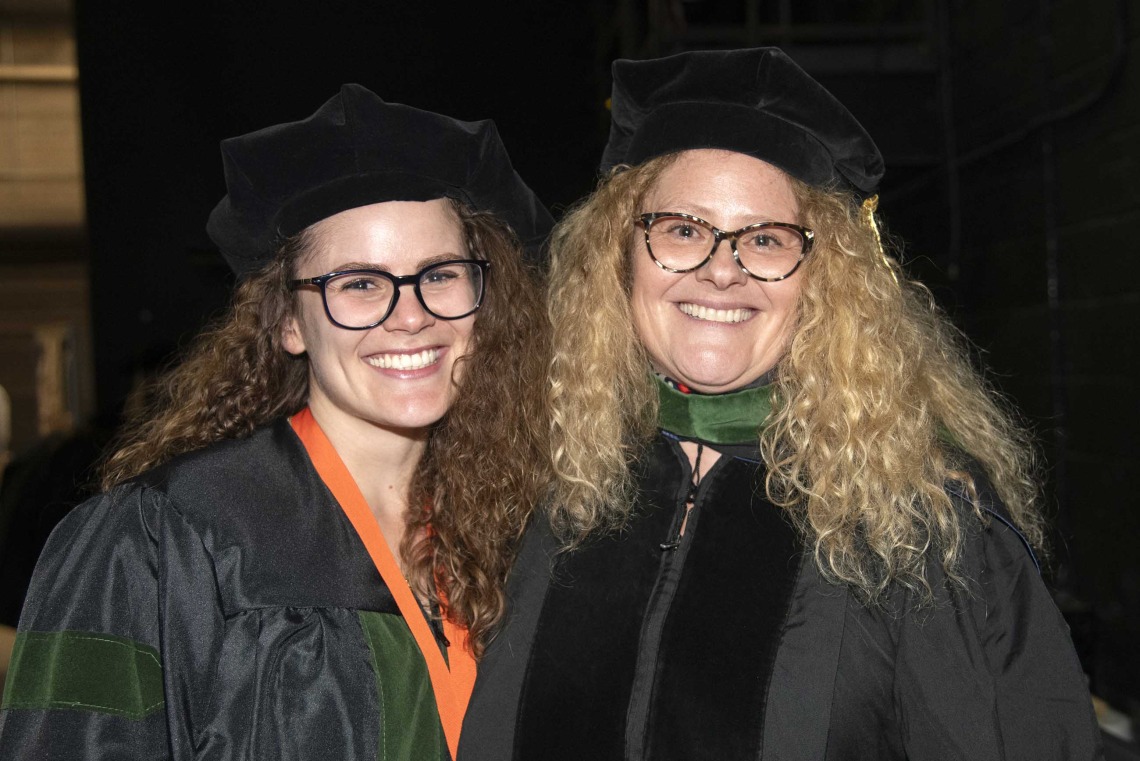 Two women, a mother and daughter, both in graduation caps and gowns, stand together, smiling, before the University of Arizona College of Medicine – Tucson commencement ceremony. 
