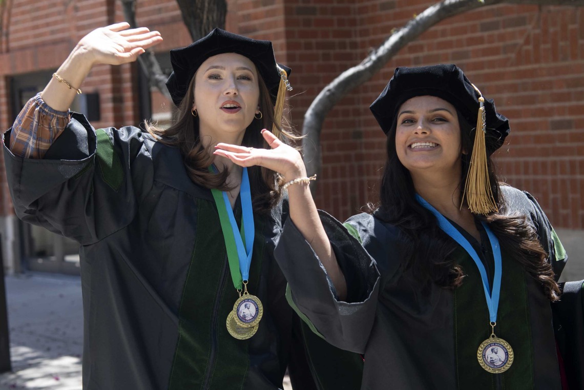 Two University of Arizona College of Medicine – Tucson students in graduation regalia wave as they walk outside on their way to their graduation ceremony.