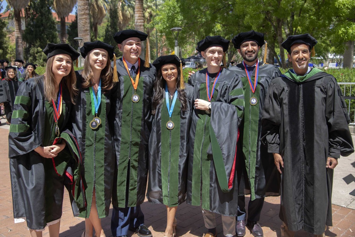 Six University of Arizona College of Medicine – Tucson students and one professor, all in graduation regalia, stand together outside before going to their commencement ceremony. 
