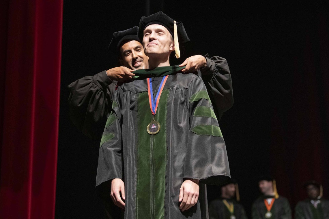 A University of Arizona College of Medicine – Tucson student dressed in a graduation cap and gown has a ceremonial hood placed over his shoulders by his faculty mentor, who is also dressed in graduation regalia.