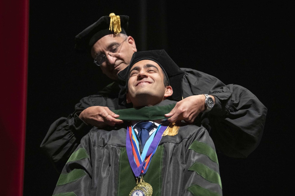 A University of Arizona College of Medicine – Tucson student dressed in a graduation cap and gown has a ceremonial hood placed over his shoulders by his father, who is also dressed in graduation regalia.