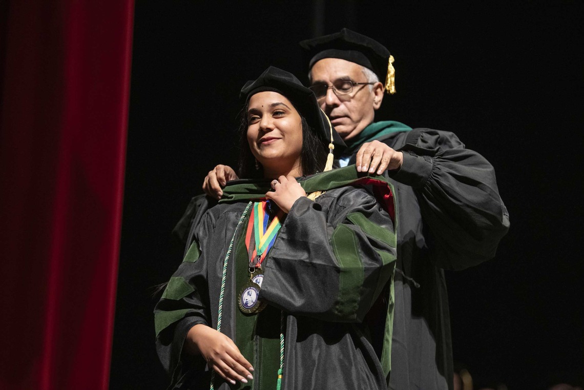 A University of Arizona College of Medicine – Tucson student dressed in a graduation cap and gown has a ceremonial hood placed over her shoulders by her father, who is also dressed in graduation regalia. 