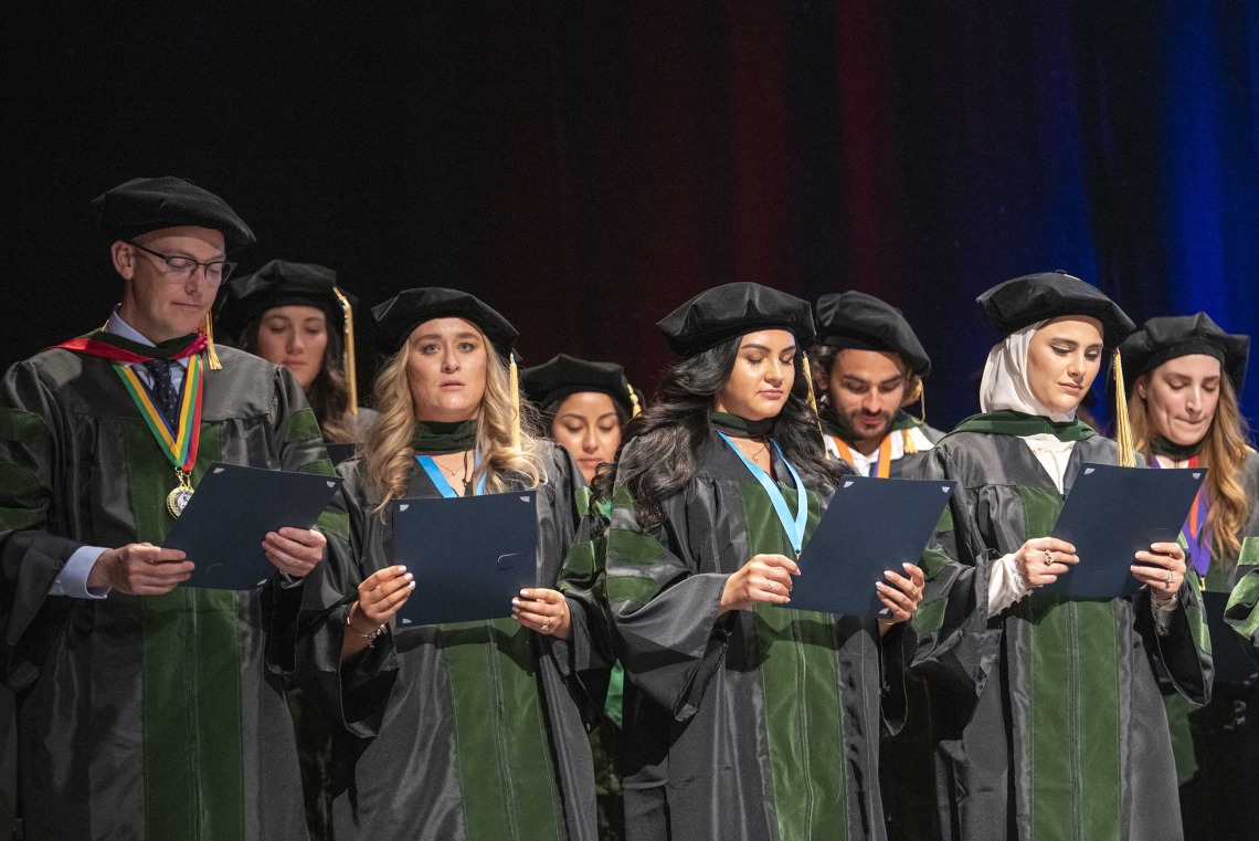 A large group of University of Arizona College of Medicine – Tucson graduates dressed in caps and gowns stand on a stage reading the Hippocratic Oath. 