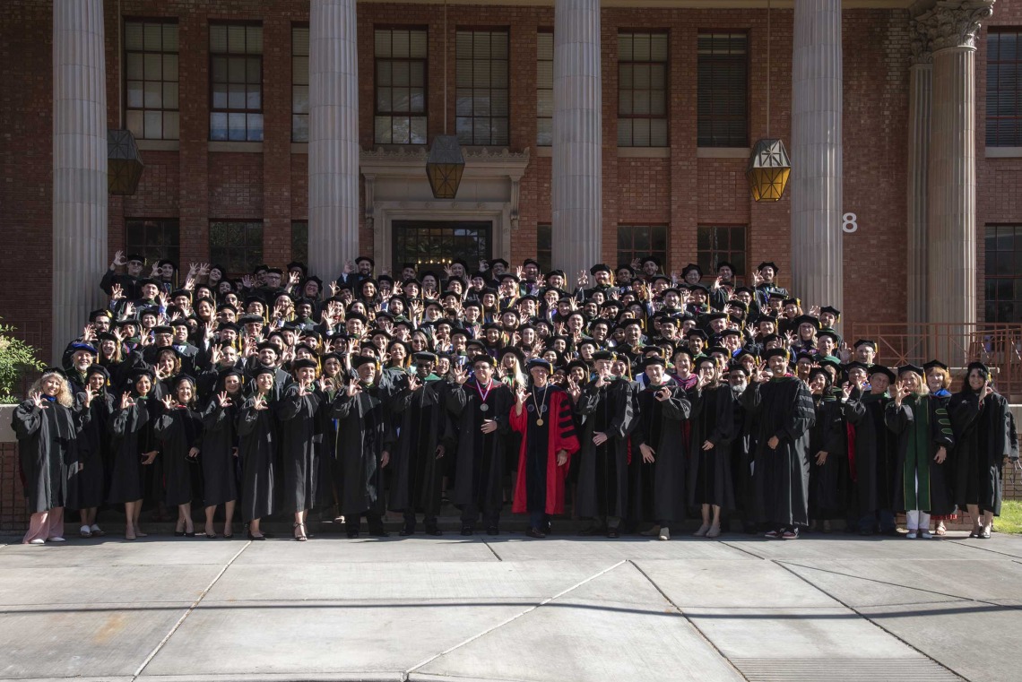 More than 100 University of Arizona College of Medicine – Tucson class of 2024 graduates and faculty stand on the stairs outside a building on the UArizona campus. All are dressed in graduation regalia. 