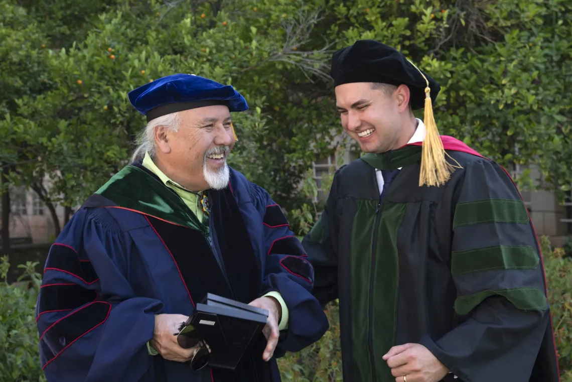 A University of Arizona College of Medicine – Tucson professor laughs with a recently graduated medical student after the commencement ceremony. Both are dressed in graduation caps and gowns. 