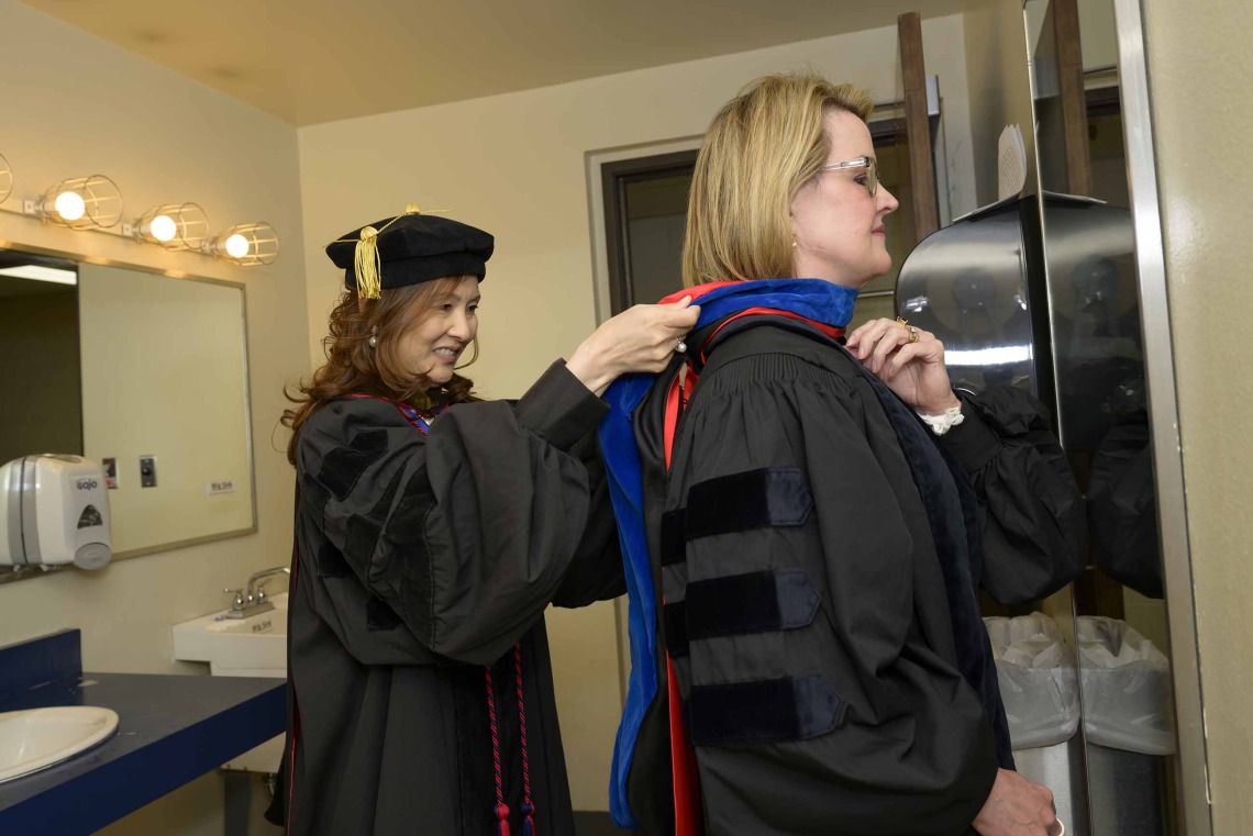 A University of Arizona R. Ken Coit College of Pharmacy faculty member dressed in graduation regalia helps another faculty member straighten her hood. 
