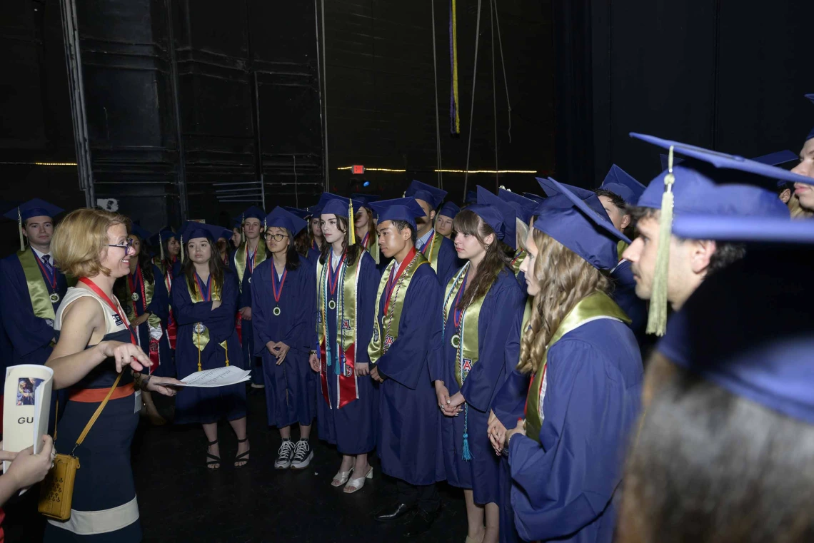 A large group of University of Arizona R. Ken Coit College of Pharmacy students wearing graduation caps and gowns stands together listening to a staff member.