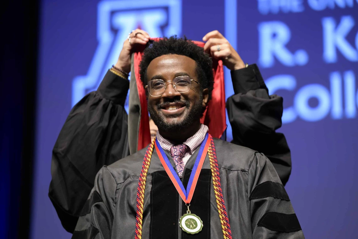 A University of Arizona R. Ken Coit College of Pharmacy student in a graduation gown smiles as a professor places a graduation hood over his head. 