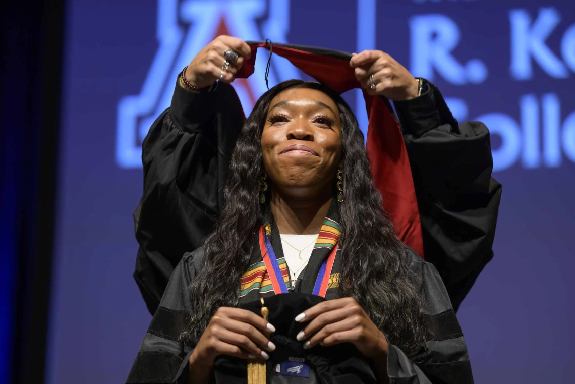 A University of Arizona R. Ken Coit College of Pharmacy student in a graduation gown smiles as a professor places a graduation hood over her head.