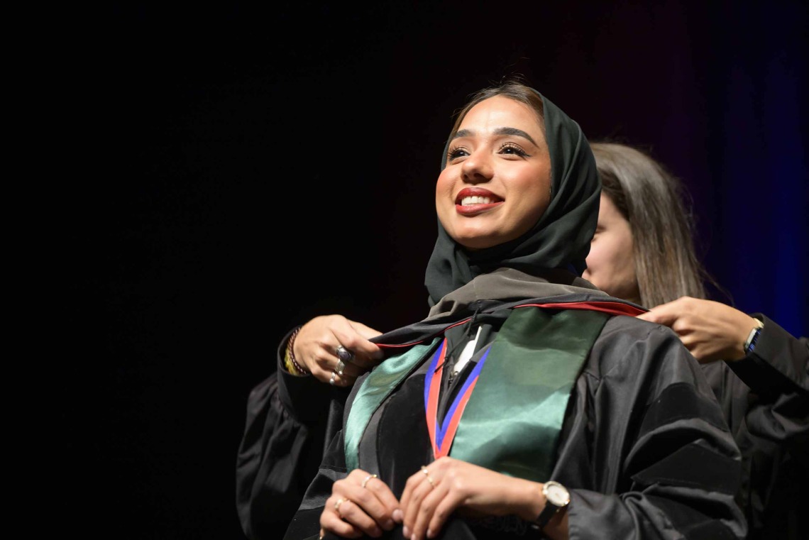 A University of Arizona R. Ken Coit College of Pharmacy Doctor of Pharmacy student wearing a graduation gown has a ceremonial hood placed over her shoulders as she smiles. 