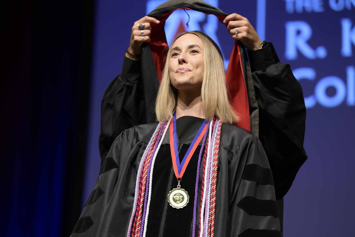 A University of Arizona R. Ken Coit College of Pharmacy student in a graduation gown smiles as a professor places a graduation hood over her head.