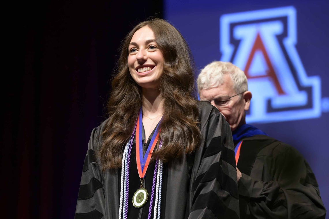 A University of Arizona R. Ken Coit College of Pharmacy student in a graduation gown smiles as a professor prepares to place a graduation hood over her head.