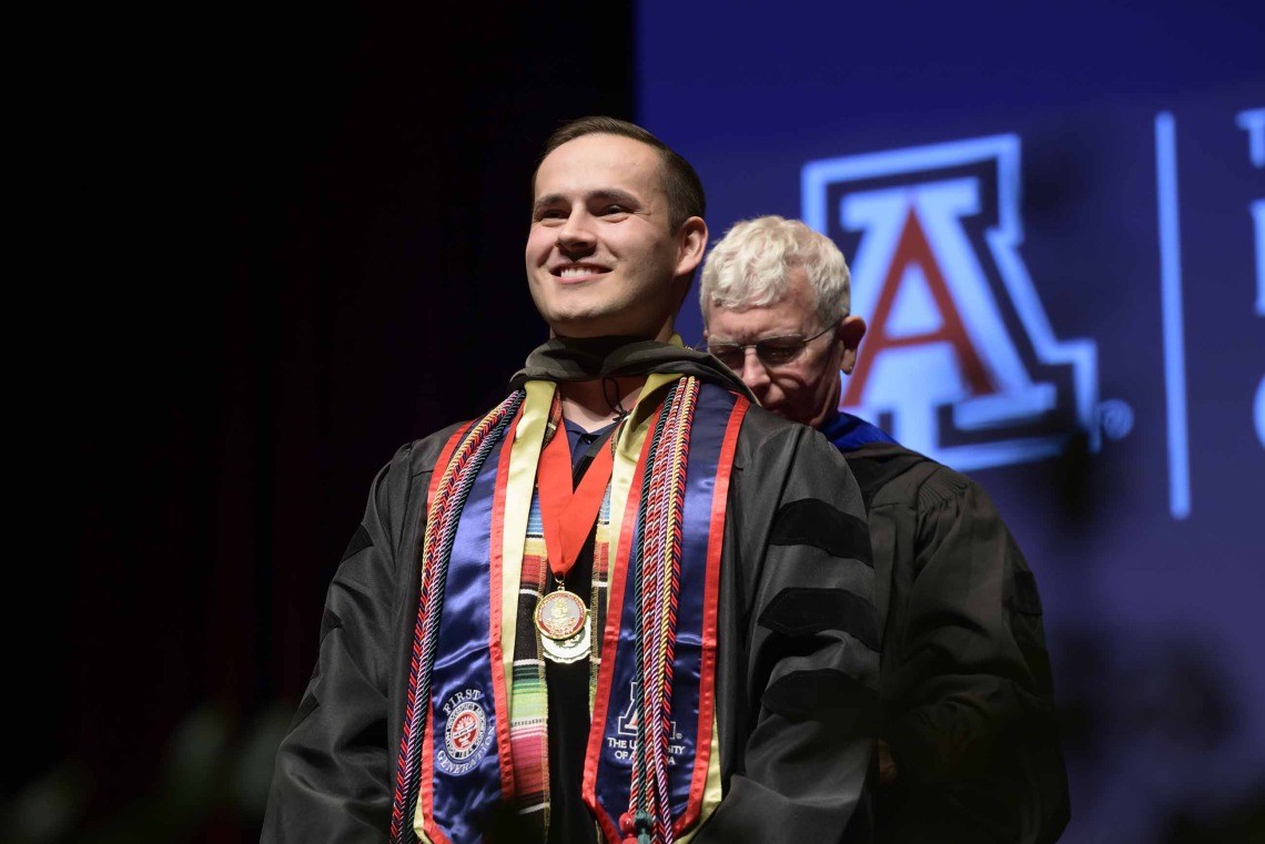 A University of Arizona R. Ken Coit College of Pharmacy student in a graduation gown smiles as a professor prepares to place a graduation hood over his head.