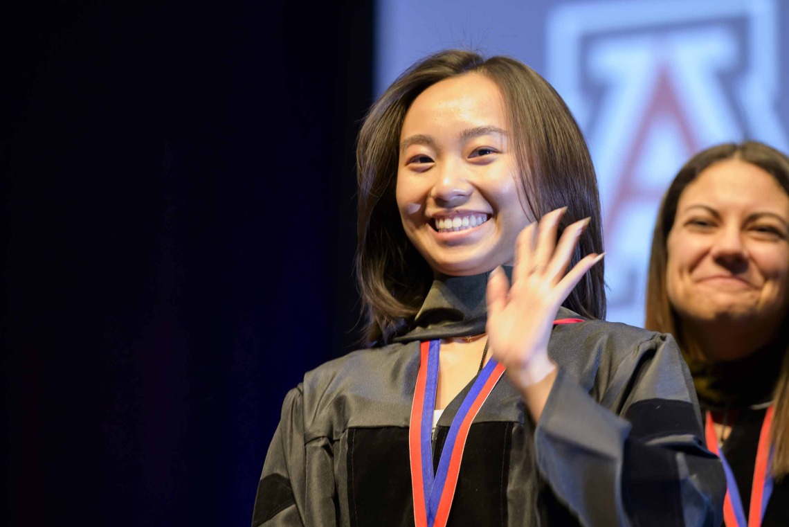A University of Arizona R. Ken Coit College of Pharmacy student dressed in a graduation gown waves and smiles as a faculty member smiles behind her.