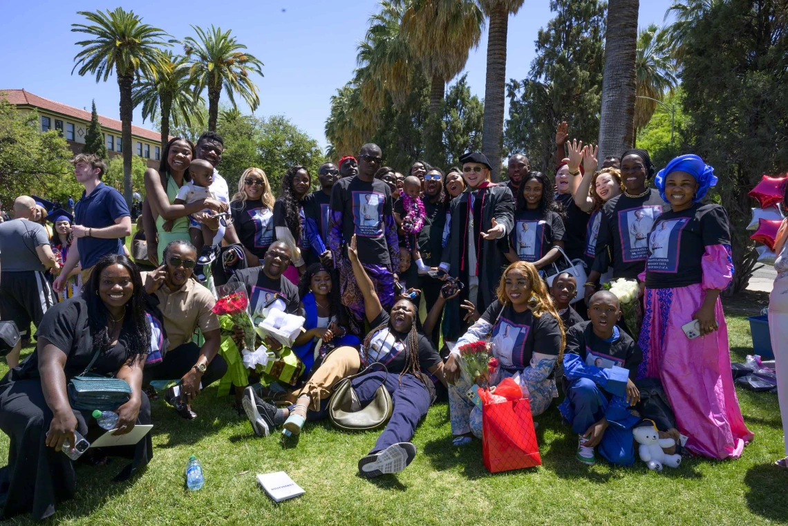 About 20 friends and family members surround a University of Arizona R. Ken Coit College of Pharmacy graduate who stands in the middle of the group wearing his graduation cap and gown. 